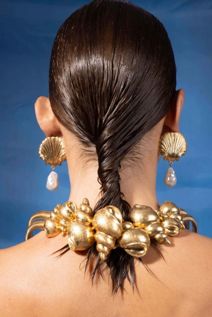 Adult female displays gold seashell jewelry featuring shell earrings with pearl drops and a large shell necklace on a blue background.