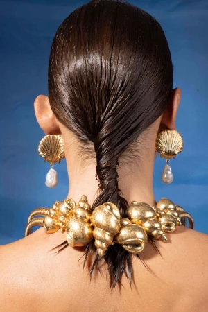 Adult female displays gold seashell jewelry featuring shell earrings with pearl drops and a large shell necklace on a blue background.
