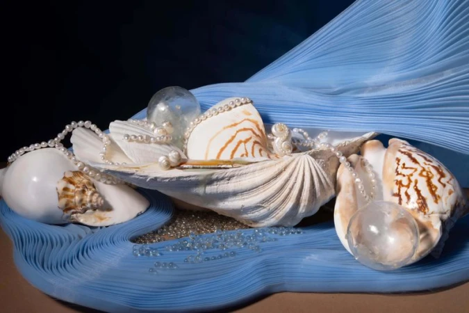 Elegant still life featuring sea shells, pearls, and glass beads on a light blue rippled cloth against a dark background.
