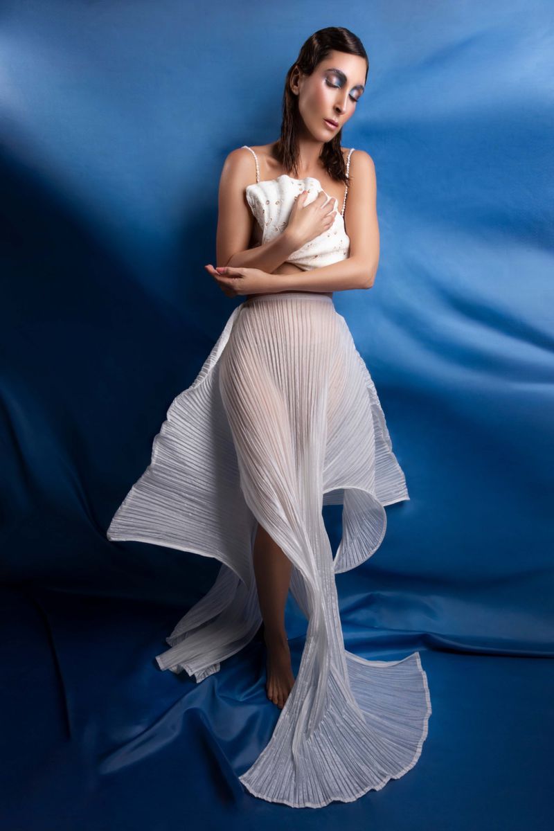 Woman in flowing white dress poses against a draped blue background, eyes closed, holding a small beaded handbag, artful fashion portrait.