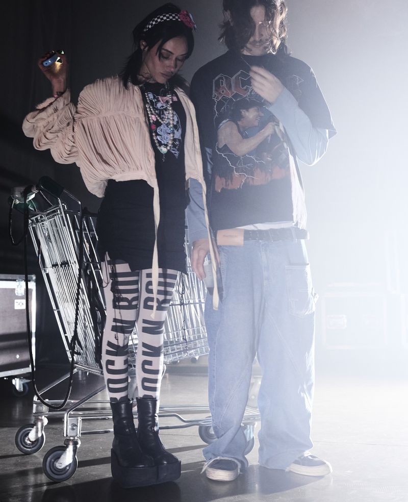 Two stylish young adults in an indoor setting with a shopping cart, wearing alternative fashion, standing in front of a bright light.