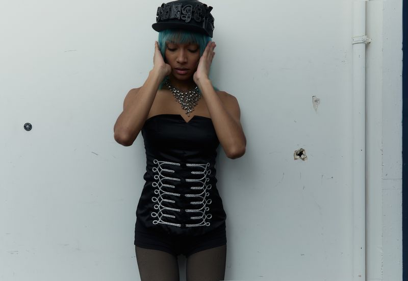 A young woman with blue hair wearing a black top and shorts poses against a white wall with one hand on either side of her head.