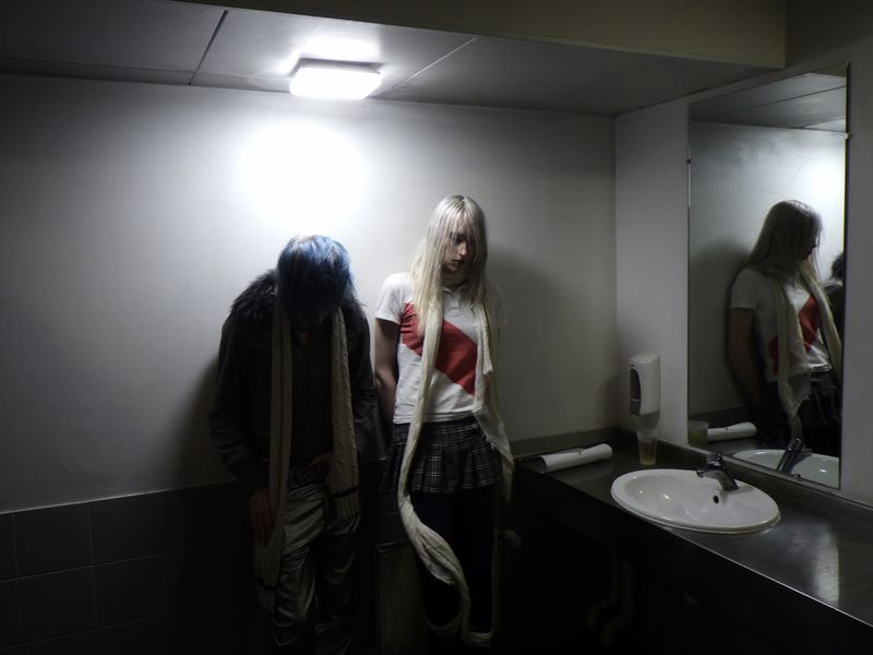 Two people with alternative fashion stand inside a bathroom with a mirror, white walls, and fluorescent lighting casting shadows.