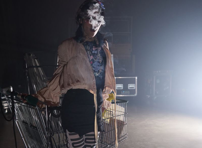 A woman in a stylish outfit pulls a shopping cart while smoking in a dark environment with stage equipment in the background.