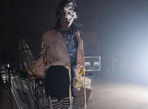 A woman in a stylish outfit pulls a shopping cart while smoking in a dark environment with stage equipment in the background.