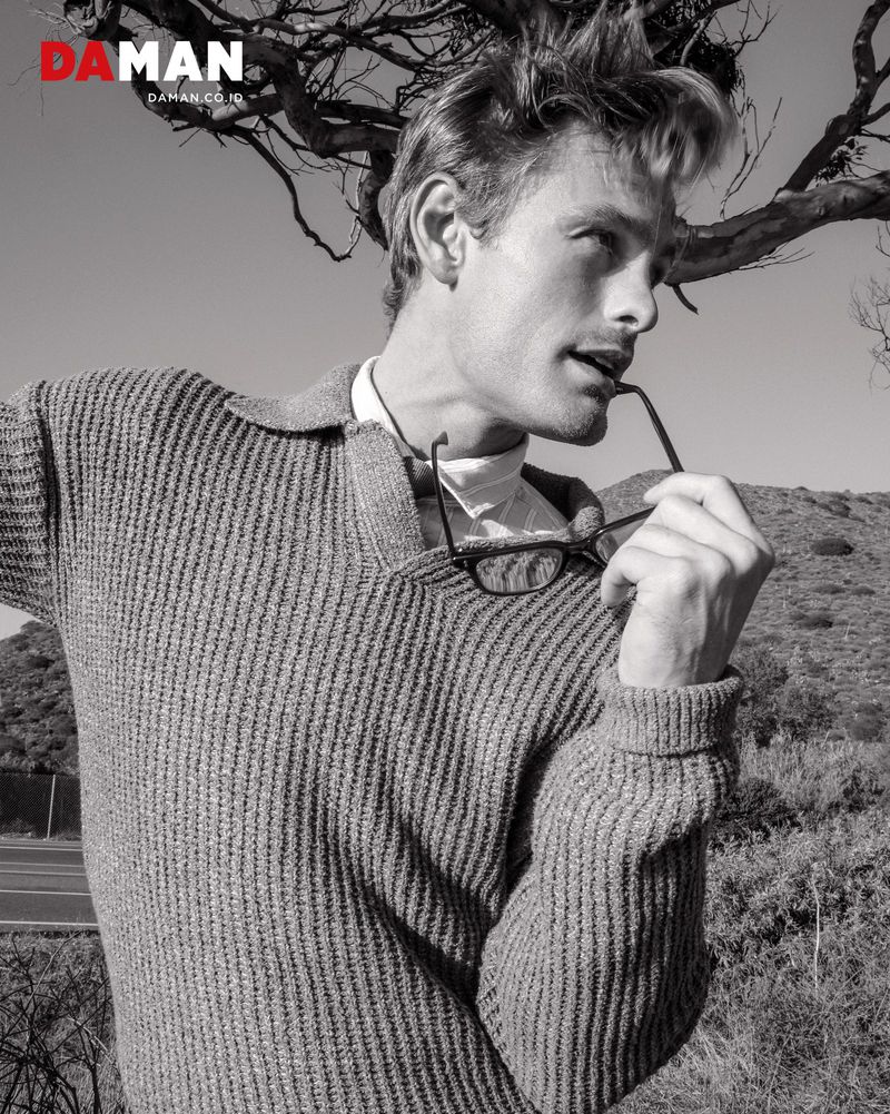 Black and white outdoor portrait of a blond adult male model in a sweater and collared shirt holding glasses by a hillside.