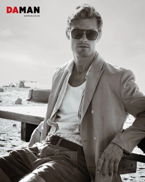 Model Noah Teicher wearing sunglasses, an unbuttoned blazer, and a white tank top seated outdoors for DA MAN Magazine.