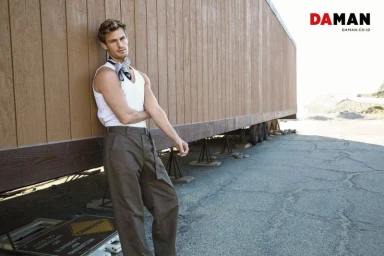 Model Noah Teicher leaning against a wooden structure in a white tank top, brown trousers, and neck scarf for DA MAN Magazine.