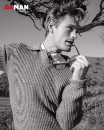 Black and white portrait of male model Noah Teicher holding glasses outdoors against a hilly background for DA MAN Magazine.