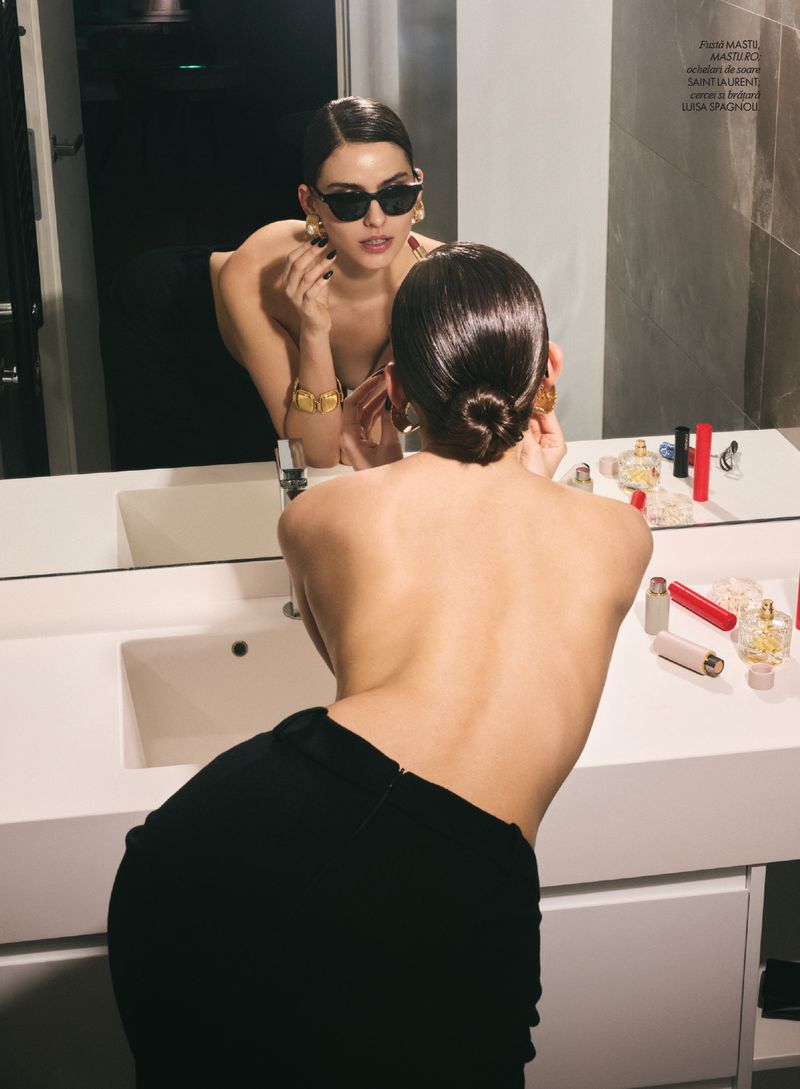 Woman in black skirt looks in a bathroom mirror, showcasing gold jewelry and makeup on the vanity in an indoor setting.