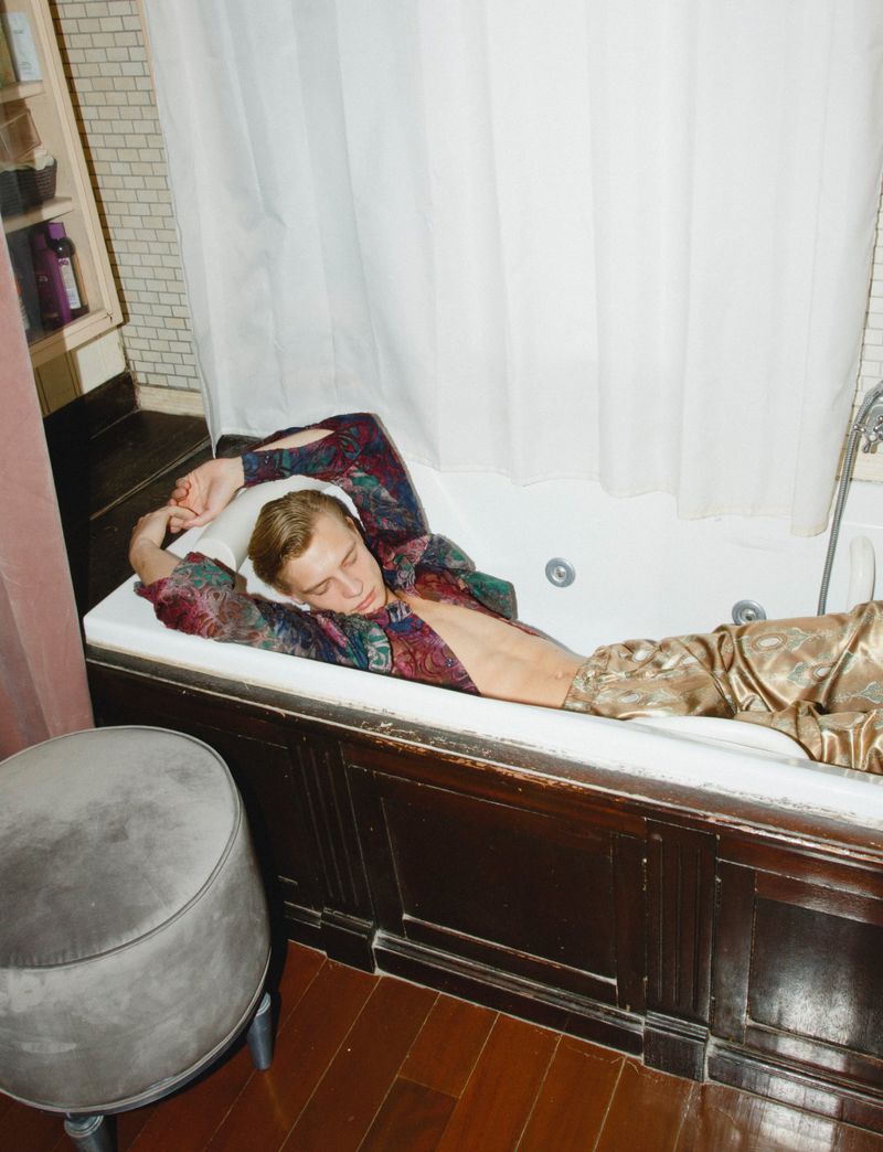 A young man relaxes in a bathtub, dressed in patterned shirt and pants, bathed in soft light against a tiled backdrop.