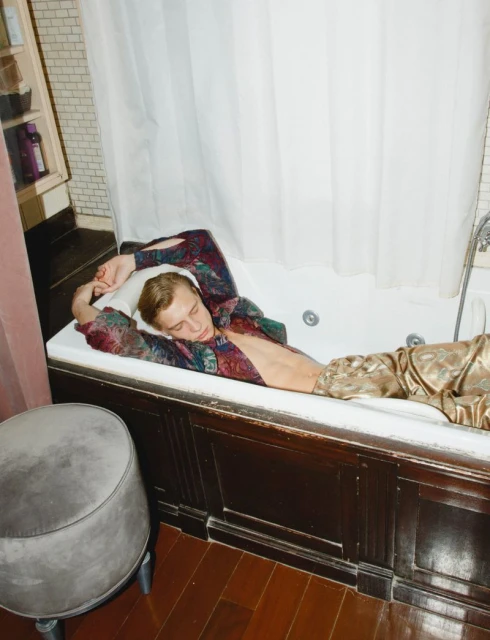 A young man relaxes in a bathtub, dressed in patterned shirt and pants, bathed in soft light against a tiled backdrop.