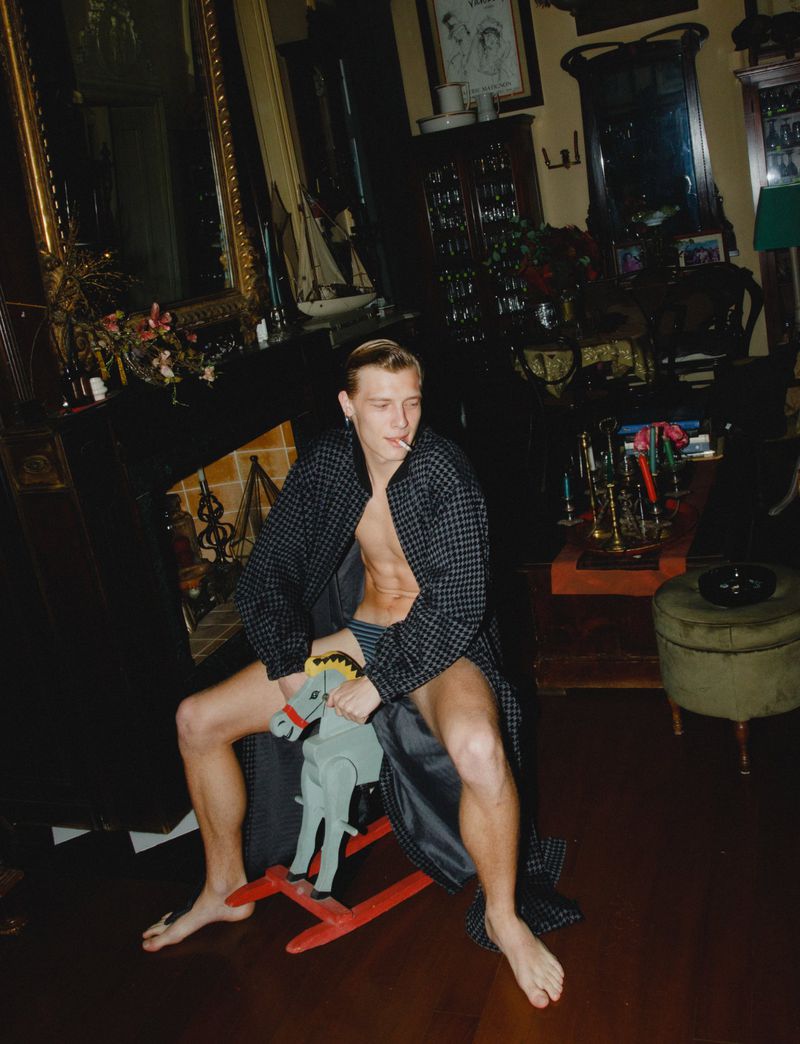 Adult male with bare chest smoking a cigarette and riding a toy rocking horse indoors, in a dark room with antique decor.
