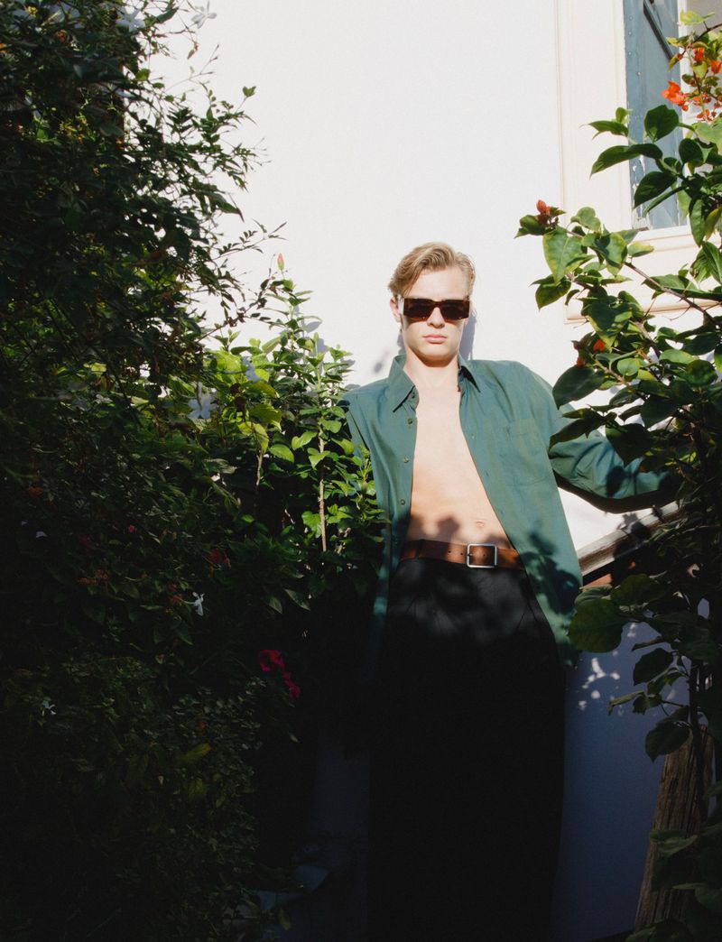 Man posing outdoors with green shirt and sunglasses, surrounded by lush greenery and bright sunlight against a white wall.