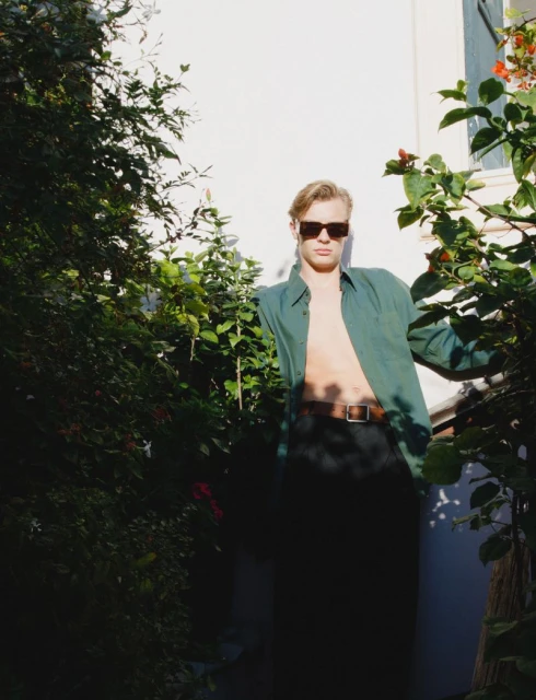 Man posing outdoors with green shirt and sunglasses, surrounded by lush greenery and bright sunlight against a white wall.