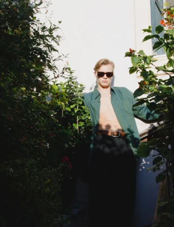 Man posing outdoors with green shirt and sunglasses, surrounded by lush greenery and bright sunlight against a white wall.