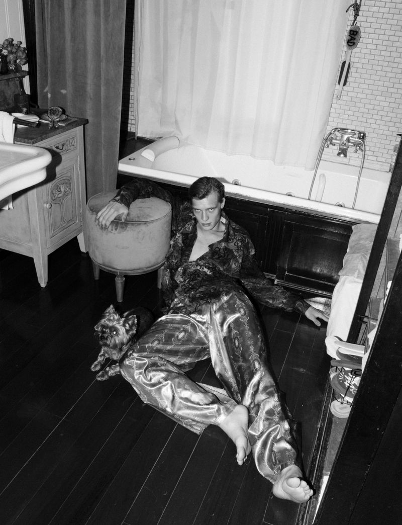 Black and white photo of a man in patterned pajamas reclining on a wood floor with a small dog, bathroom fixtures in background.