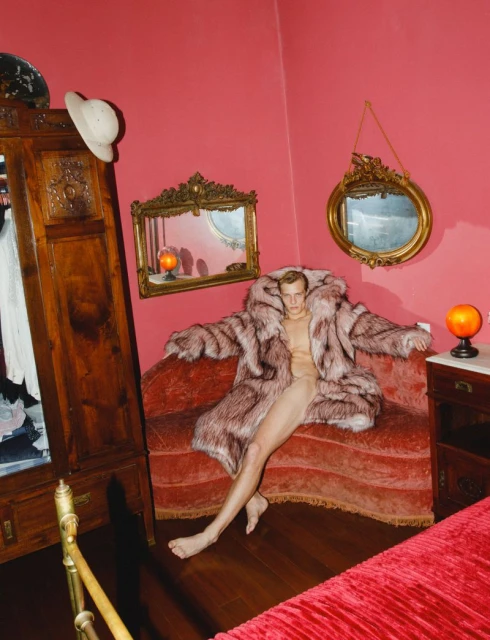 Nude adult male wearing a fur coat sits on a red sofa in an indoor setting with red walls, wardrobe and lamps.