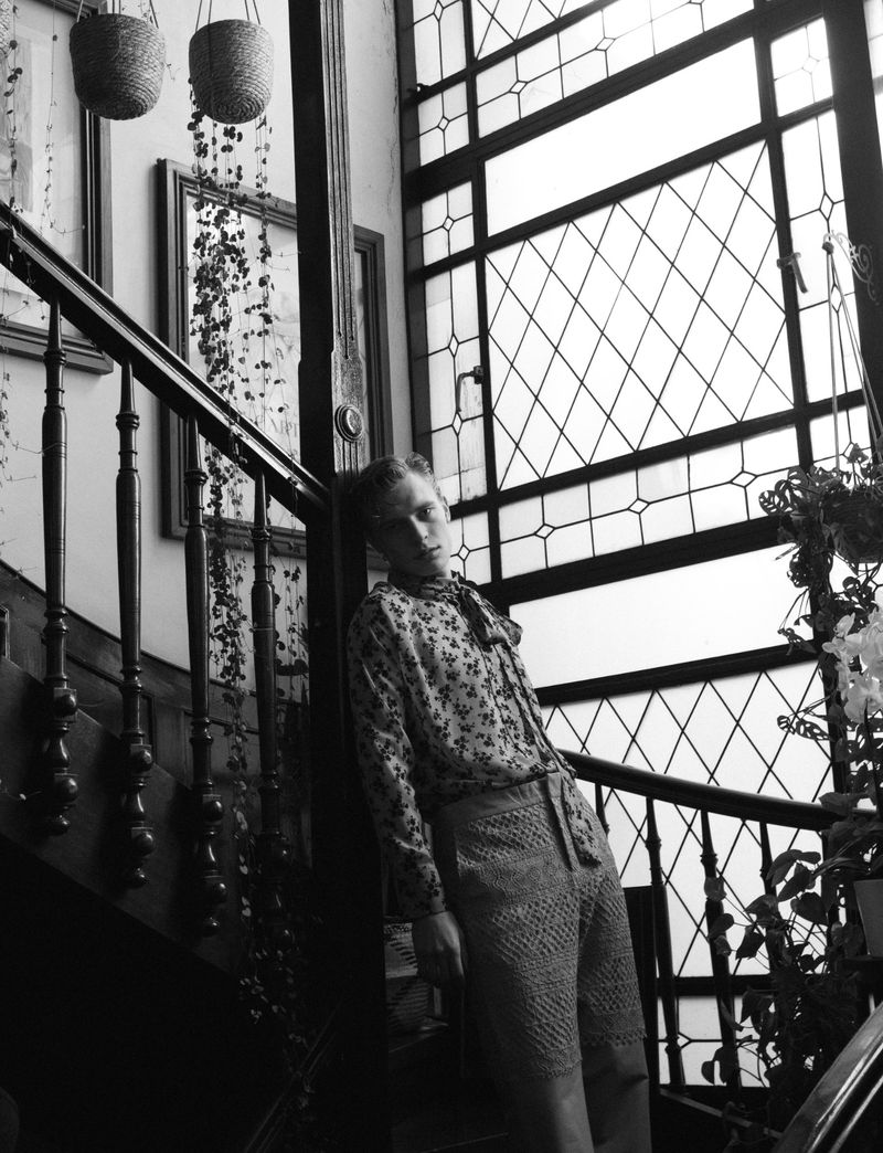 Black and white photo of a woman leaning against a wooden staircase indoors with a diamond-patterned window and plants.