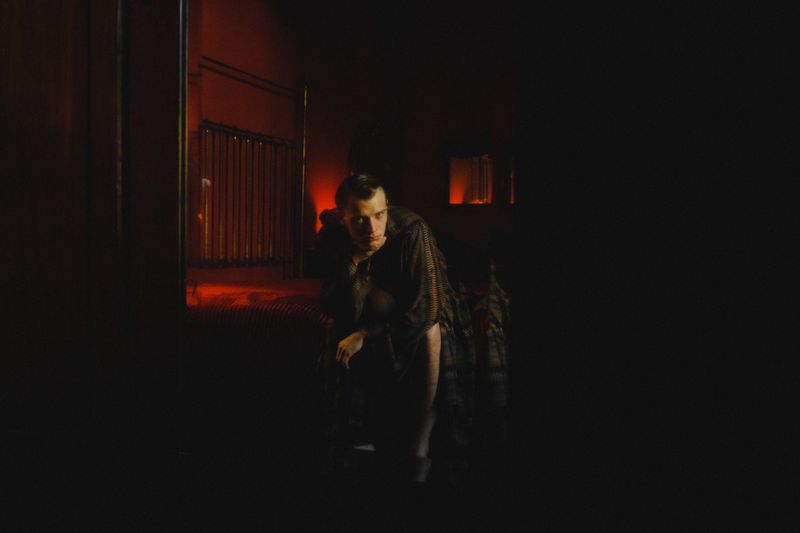 Moody portrait of a man kneeling in a dark room with dim red lighting and shadows, creating a cinematic atmosphere and intriguing composition.