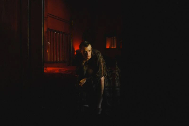 Moody portrait of a man kneeling in a dark room with dim red lighting and shadows, creating a cinematic atmosphere and intriguing composition.