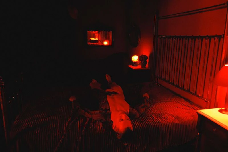 Adult lying on a red striped bed in a dark room with a mirror and a lamp on a nightstand, all illuminated by red light.