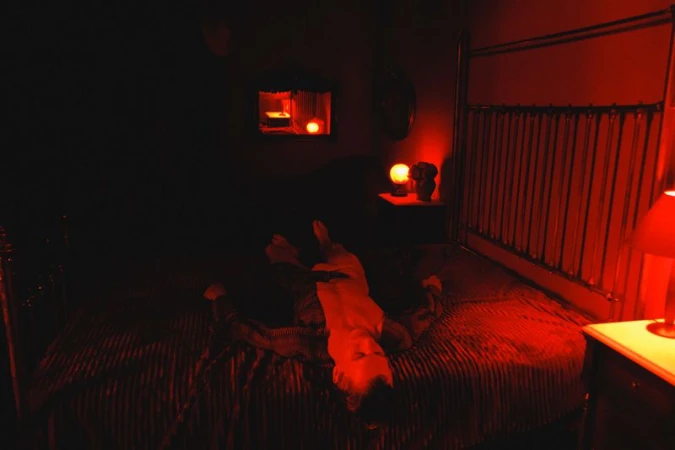 Adult lying on a red striped bed in a dark room with a mirror and a lamp on a nightstand, all illuminated by red light.