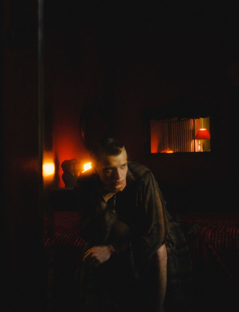 Indoor portrait of a fair-skinned adult male with short hair, wearing a patterned shirt in a room with dark red walls and dim, warm lighting.