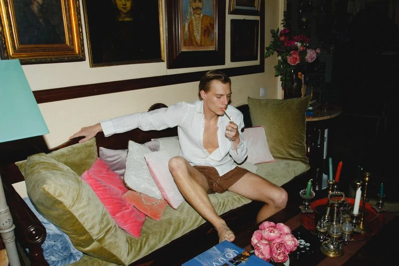 Man relaxes on a green velvet couch surrounded by decorative pillows, smoking with artwork and floral arrangements in the background.