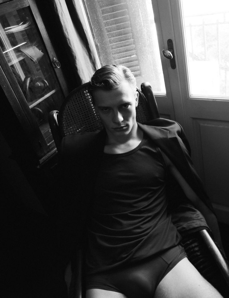 Black-and-white shot of a blond man in underwear and a blazer, seated in a wicker chair in front of a window with dark wood cabinets.
