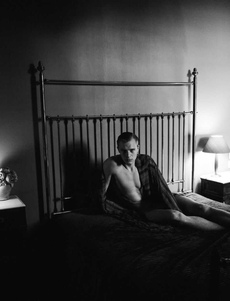 Monochrome portrait of a young man draped in a blanket seated on a metal frame bed in a dim bedroom.