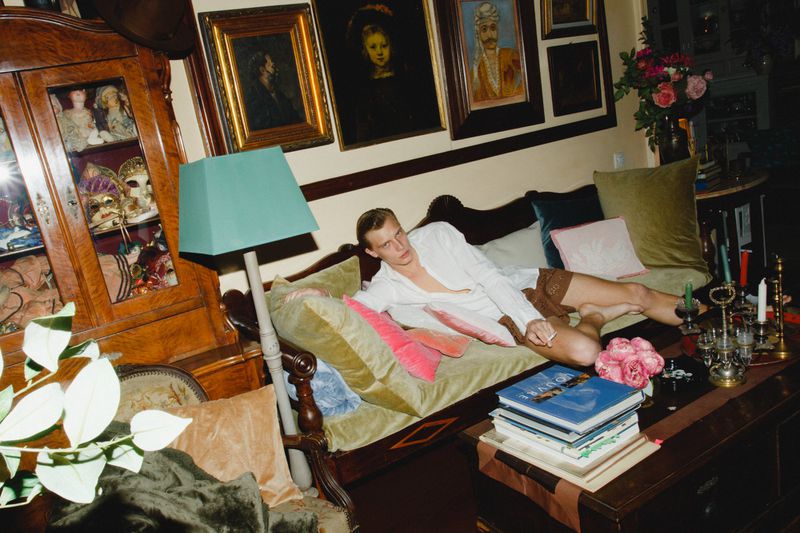 Adult male lounging on a vintage velvet sofa in an opulent interior with antique furniture, artwork, flowers, and a stacked coffee table.