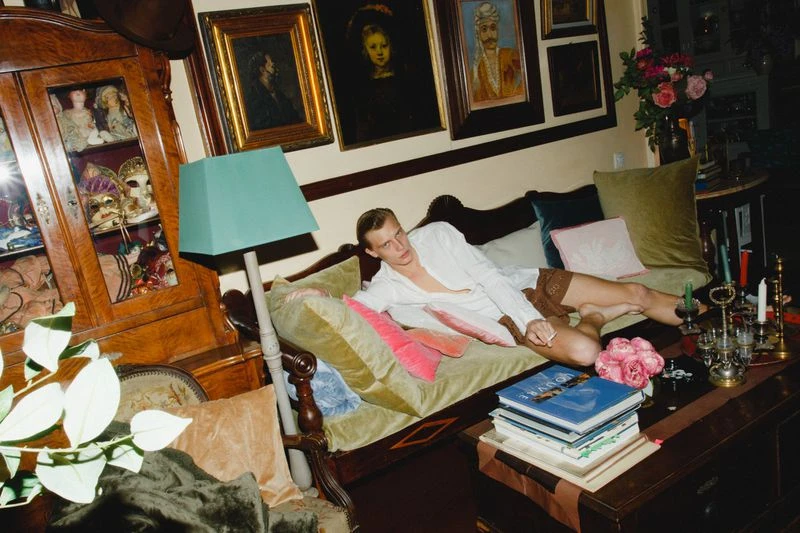 Adult male lounging on a vintage velvet sofa in an opulent interior with antique furniture, artwork, flowers, and a stacked coffee table.