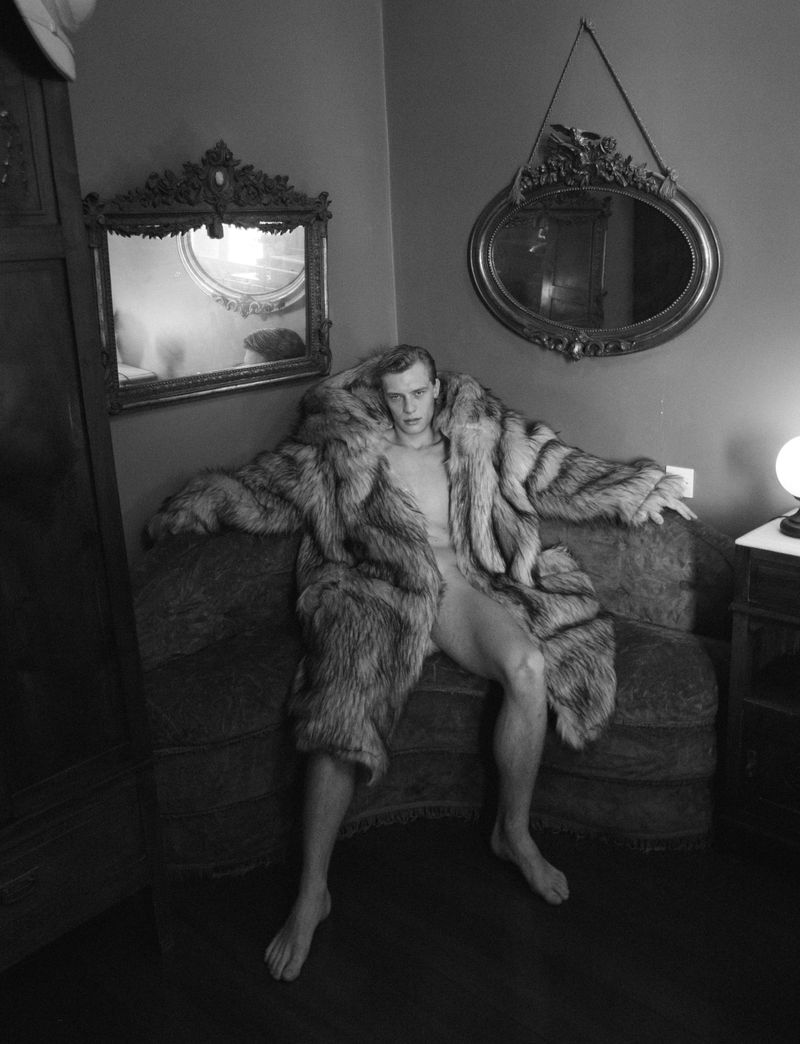 Black and white portrait of a man reclining in a fur coat on a settee, framed by vintage mirrors, in a dimly lit room.