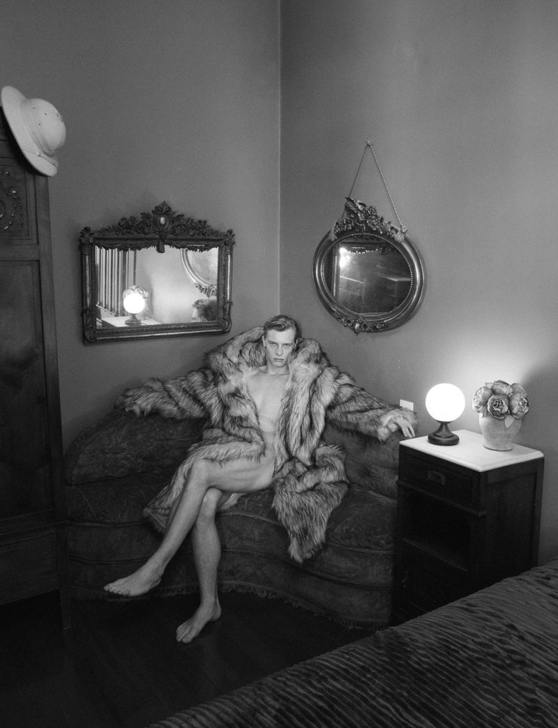 Monochrome photograph of a man seated on a sofa wearing an open oversized fur coat in a vintage interior with ornate mirrors.