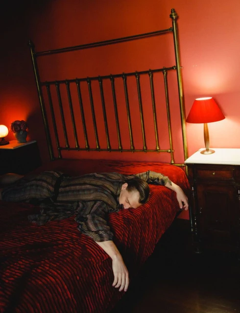 Adult male lying on a red bed in a bedroom interior with a gold bed frame and a nightstand with a red lamp and sphere lamp.