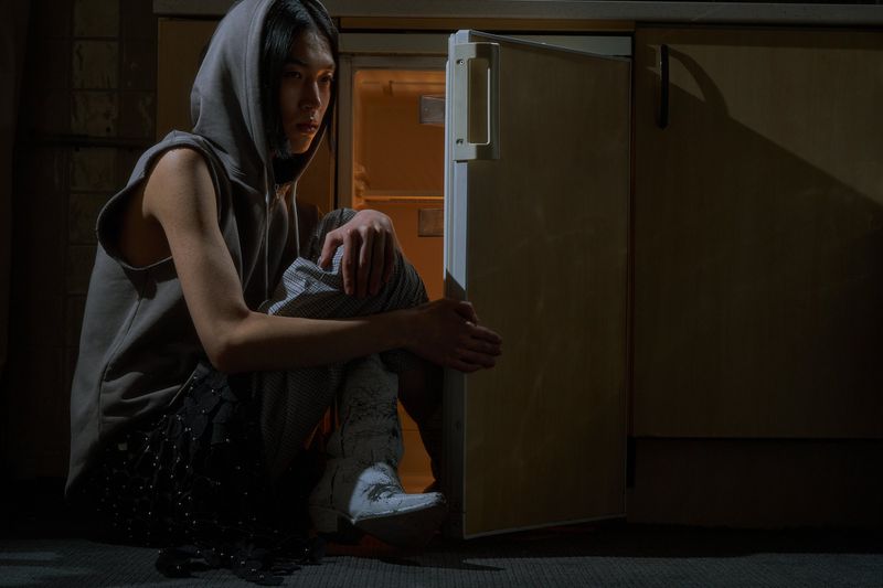 Young adult sits on the floor near a refrigerator wearing a sleeveless hoodie, patterned pants, and light-colored boots in an indoor setting.
