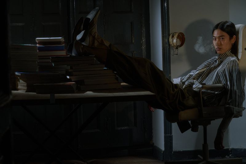 Adult male sitting in an office chair with his feet on a table covered in books, wearing a striped shirt and brown pants.