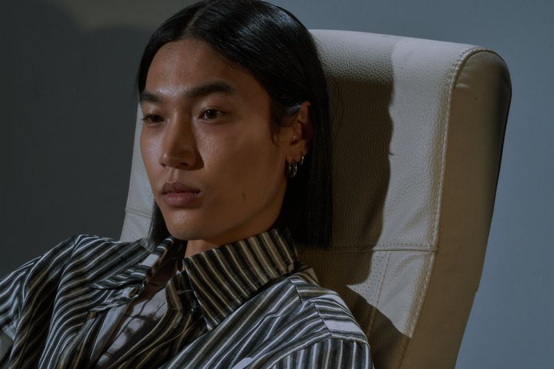 Close up portrait of an adult male wearing a vertically striped shirt, seated on a chair in an indoor studio environment.