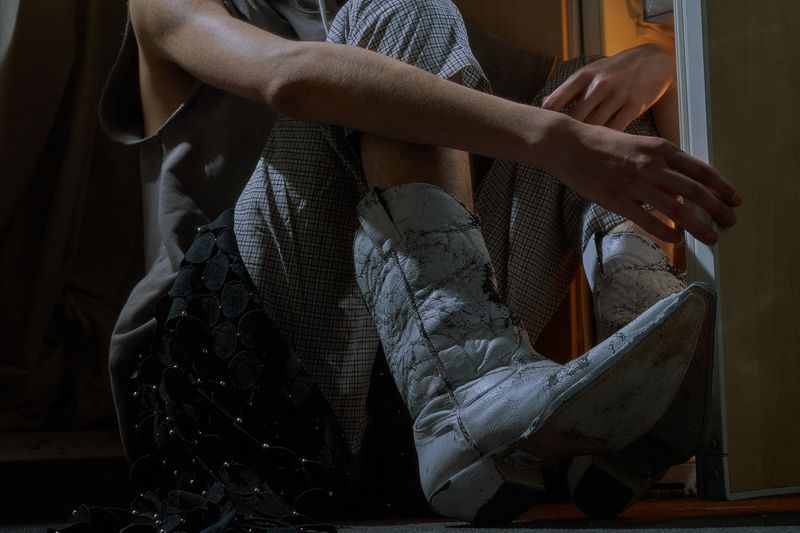 Person wearing cowboy boots and patterned pants sits indoors near a doorway in dim lighting, creating a casual and somewhat edgy style.