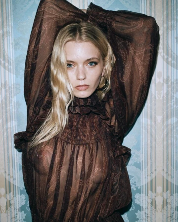 A blonde woman wearing a sheer brown patterned top posing against blue striped wallpaper