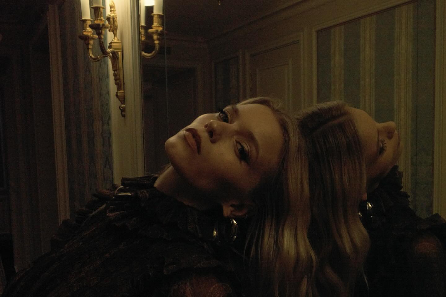Indoor portrait of a fair-skinned woman reflected in a mirror. She's wearing a lace top and gold hoop earrings, posing with head tilted back.