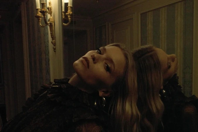 Indoor portrait of a fair-skinned woman reflected in a mirror. She's wearing a lace top and gold hoop earrings, posing with head tilted back.