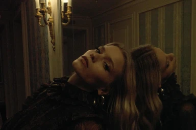 Blonde woman in dark ruffled blouse leaning against a mirror in a dimly lit room with an ornate wall sconce.