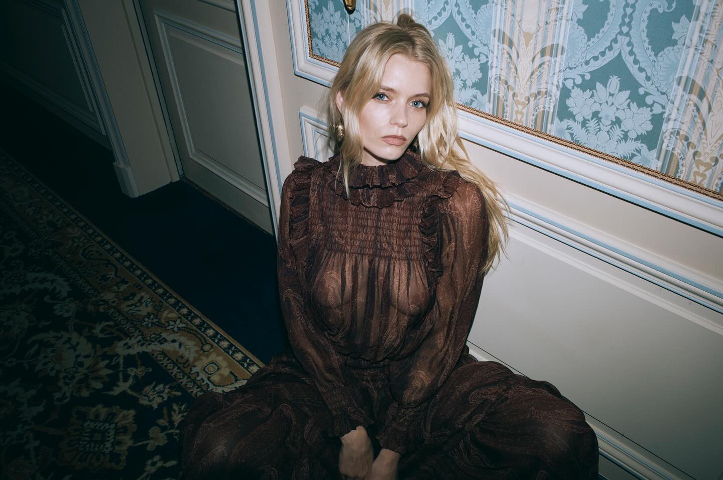 A young blond woman sits on an ornate carpet wearing a brown paisley maxi dress with ruffled neckline in an indoor setting.