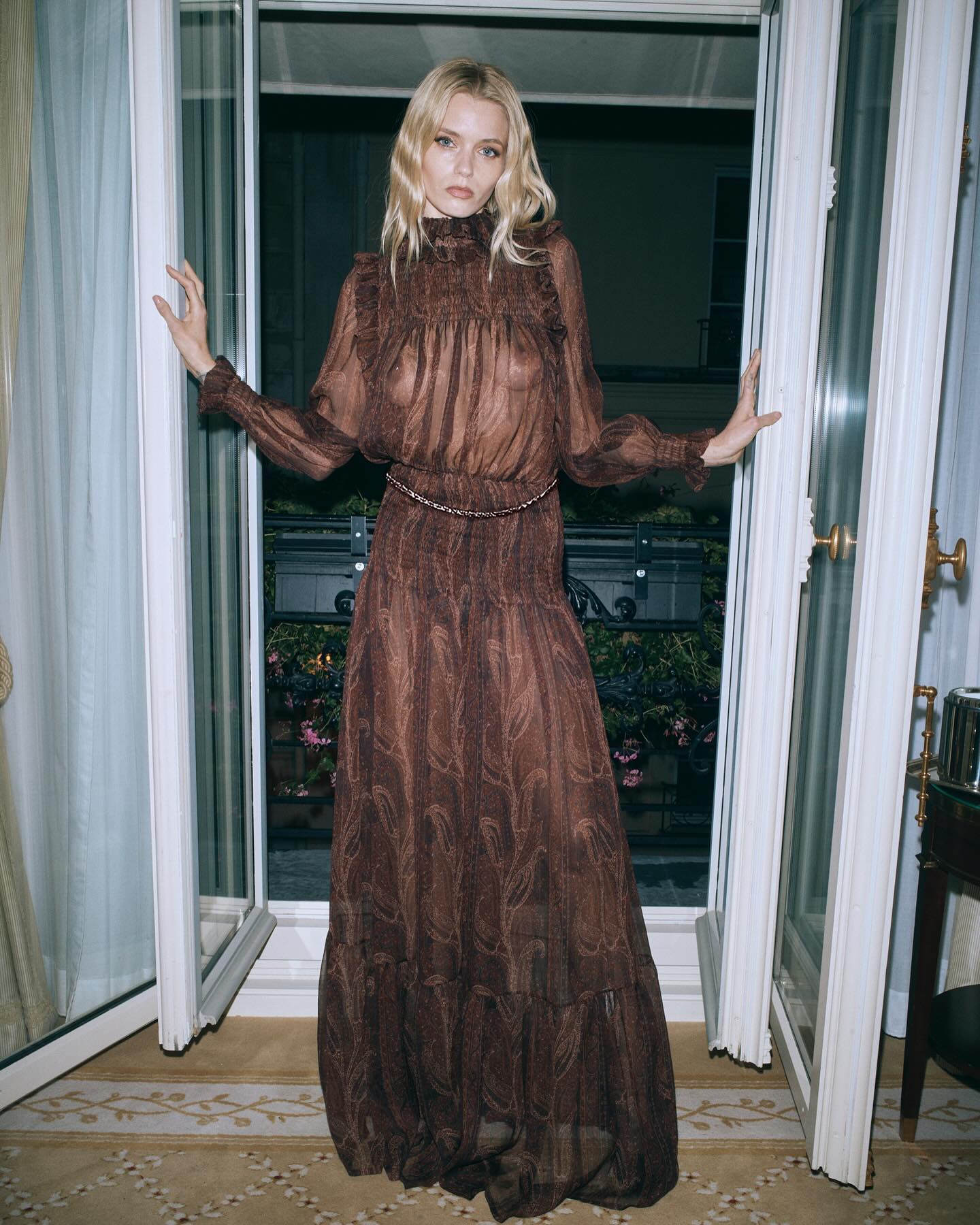 A fair-skinned woman poses in a long brown paisley dress and stands between white french doors in an indoor setting with dark lighting.