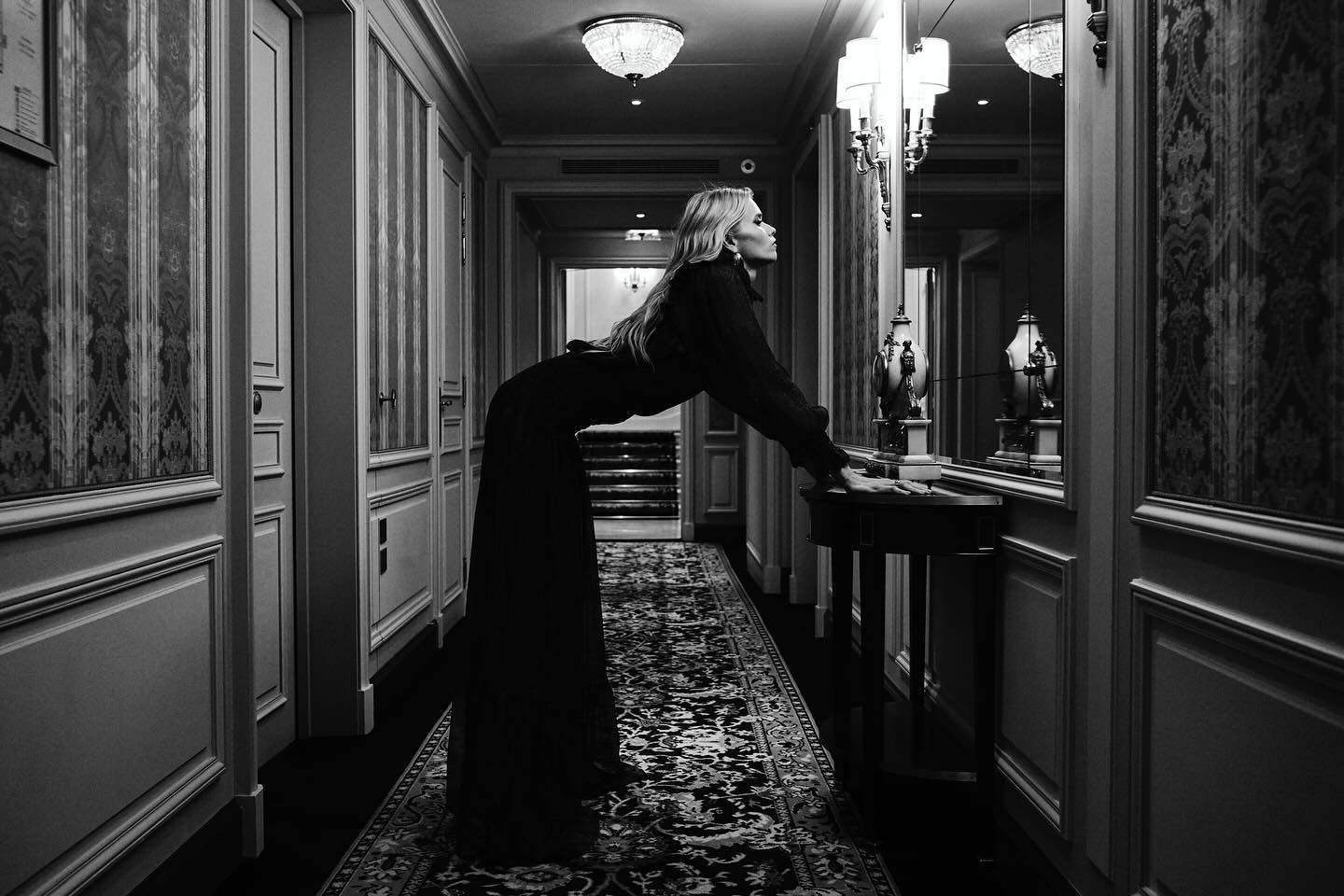 A woman in a long black dress leans on a dark wood table in a dimly lit hallway with ornate wallpaper and ceiling lights.