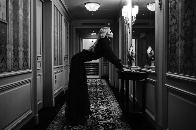 A woman in a long black dress leans on a dark wood table in a dimly lit hallway with ornate wallpaper and ceiling lights.