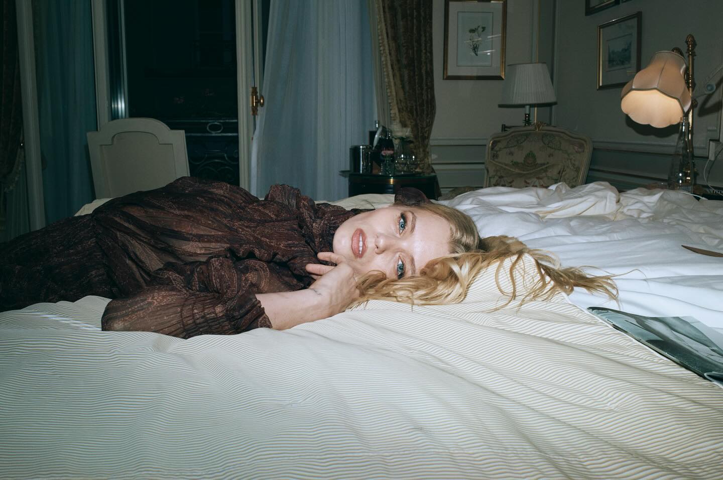 Blonde woman wearing a dark patterned dress lying on a bed in a hotel room