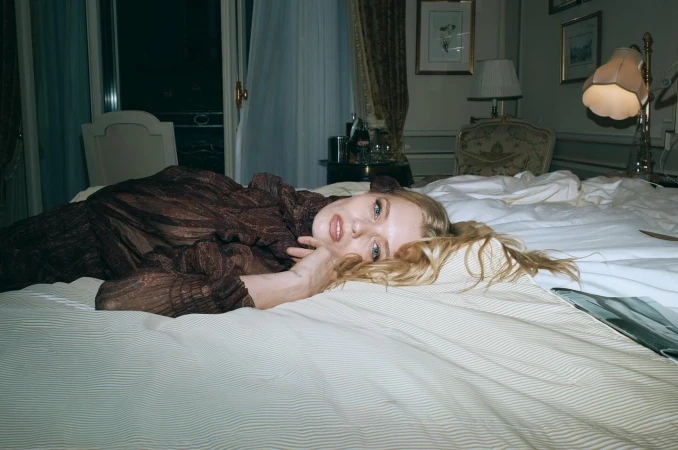 Blonde woman wearing a dark patterned dress lying on a bed in a hotel room